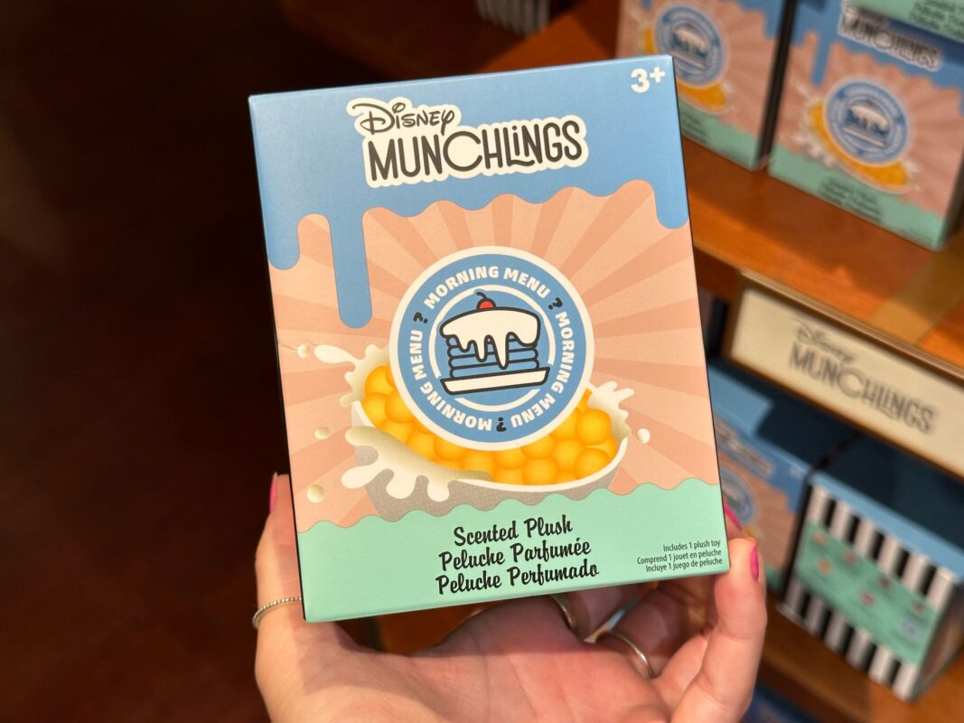 A person holds a "Disney Munchlings" scented plush toy box which features a waffle and syrup design. The box is labeled for ages 3 and up and placed among other similar boxes on a store shelf.