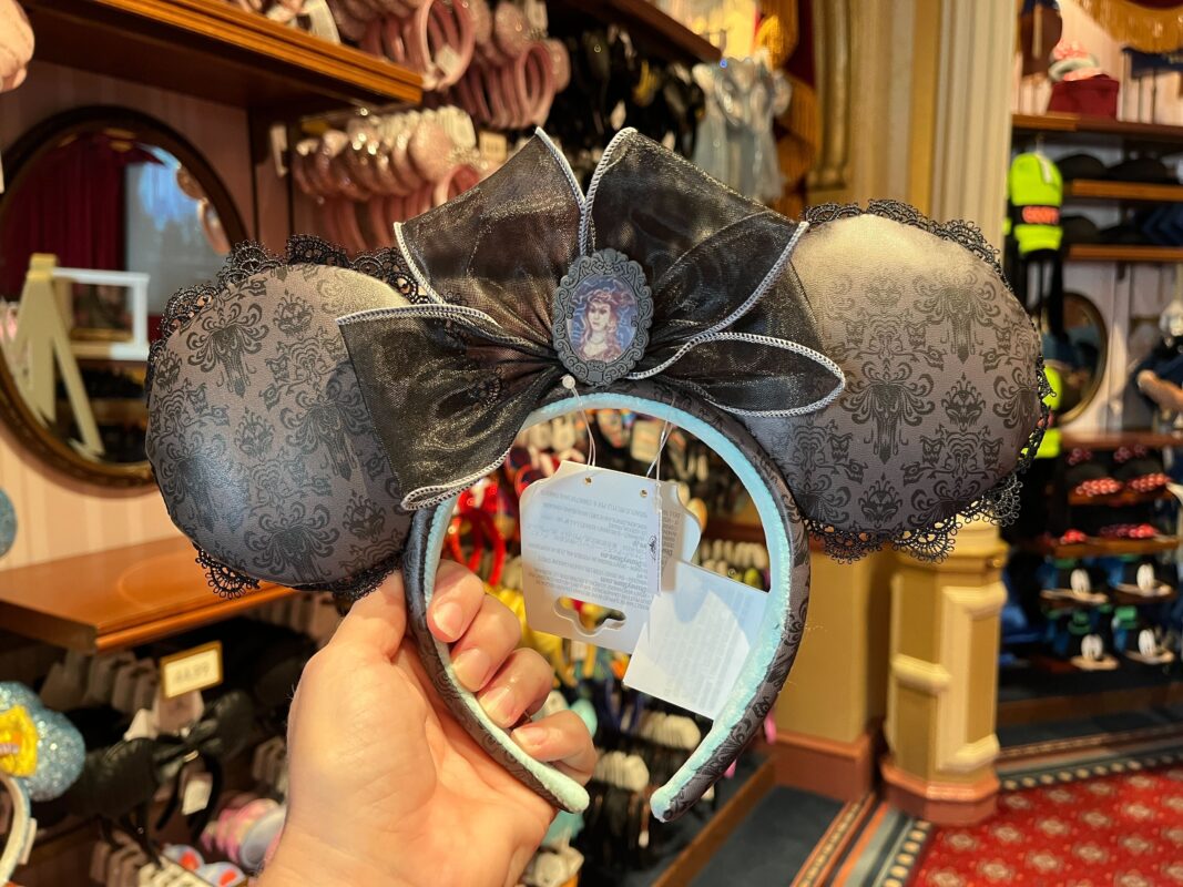 A person holds up a pair of decorative Mickey Mouse ears headband in a store, featuring a black lace pattern, a large bow, and a central cameo design. Shelves displaying various headbands are in the background.