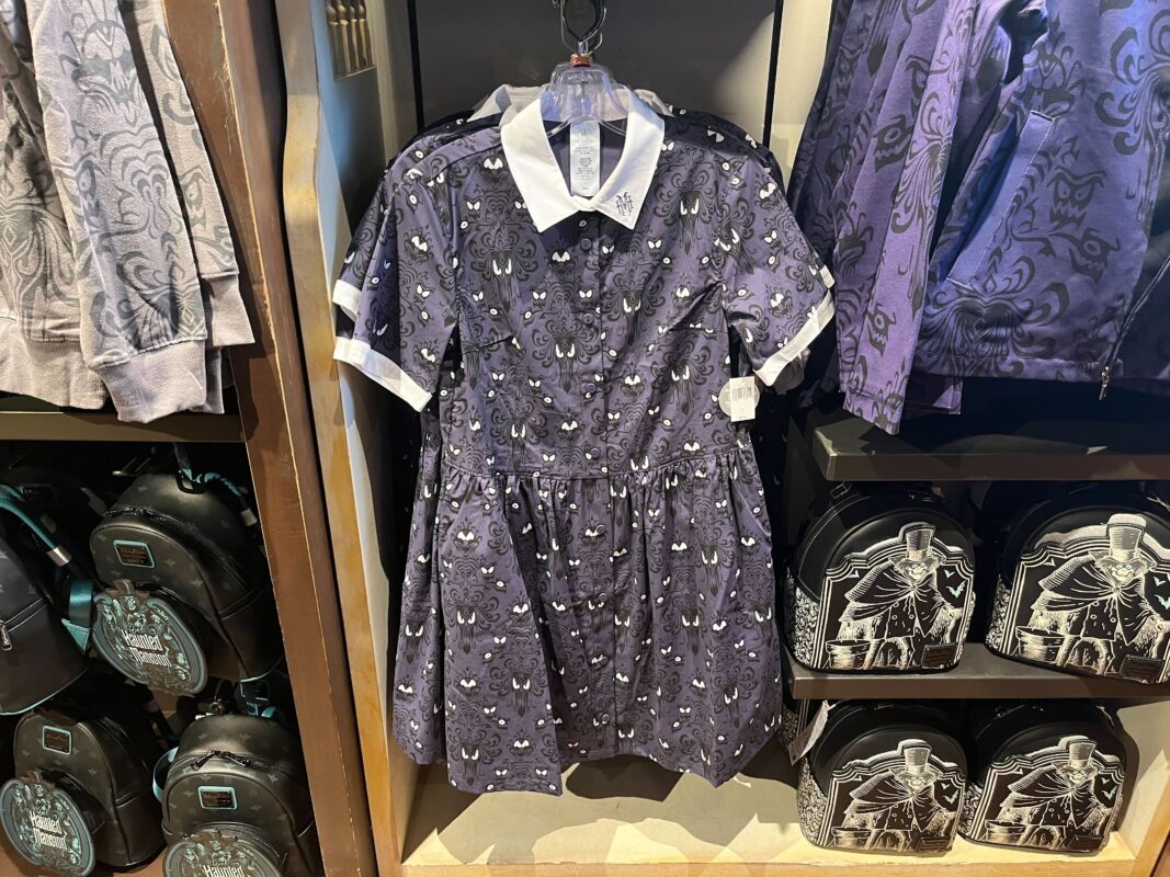 A purple dress with a white collar featuring a pattern of small, white, ghost-like faces hangs on a rack in a store. Nearby, matching backpacks and jackets are displayed.