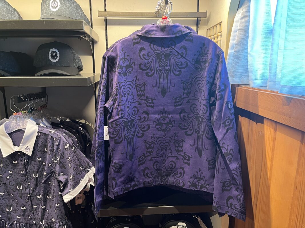 A purple jacket with black ornate patterns hangs in a clothing store. Nearby are racks with hats and dresses with similar patterns.