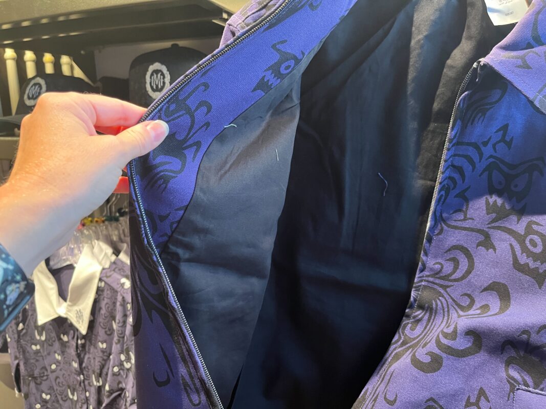 A person holds open a purple jacket adorned with black intricate patterns, revealing its plain black inner lining. Additional garments with similar print hang in the background.