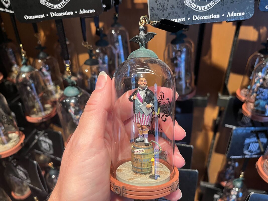 A hand holding a glass ornament featuring a miniature figure of a man with a mustache, striped pants, and a top hat, standing on a barrel. Other similar ornaments are displayed in the background.