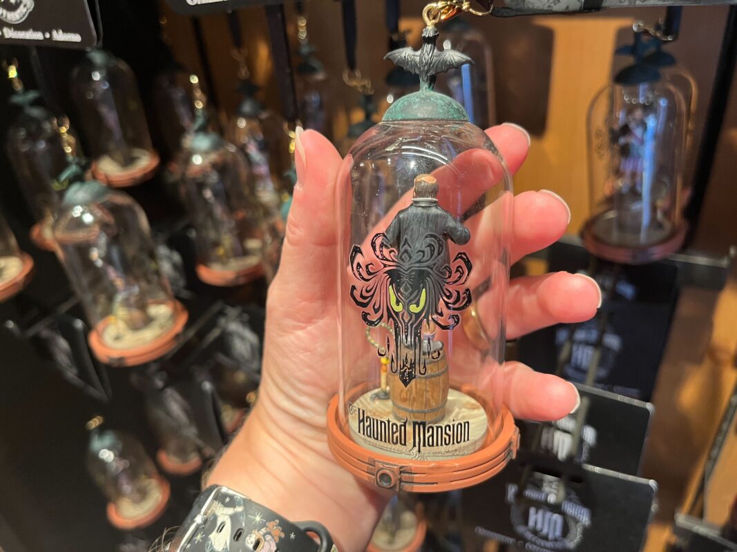 A hand holds a Haunted Mansion-themed ornament encased in a glass dome. The ornament features a figure standing on a pillar with a bat on top. Multiple similar ornaments are displayed in the background.