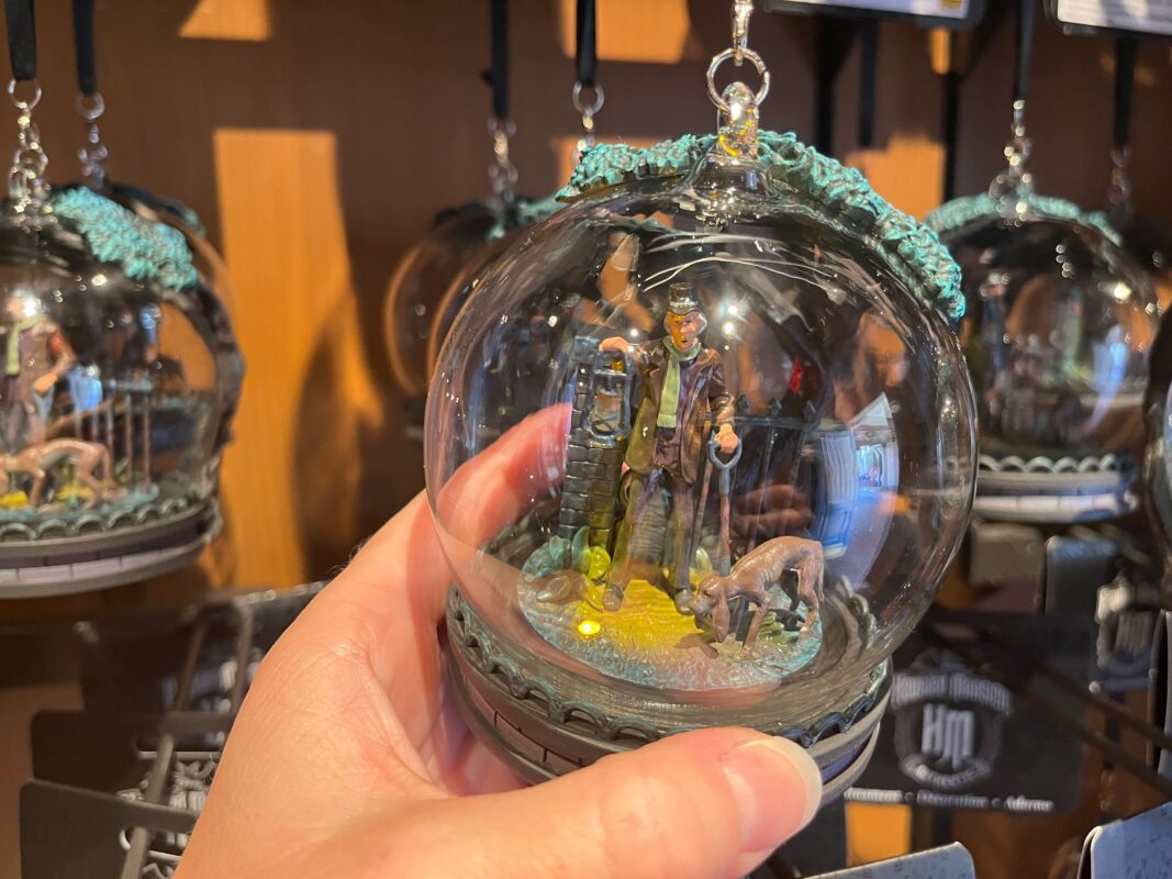 A hand holding a clear glass ornament depicting a man holding a lantern next to a crouched figure in a dim environment. Other similar ornaments are displayed in the background.