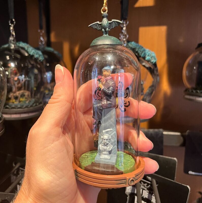 A hand holds a glass dome containing a miniature figurine of a man standing on a pedestal. Other similar domes with different figures hang in the background.
