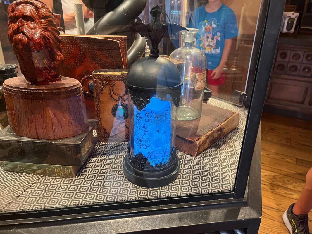 A display case holds various artifacts including a glowing blue lantern, vintage books, and a wooden carved face. A person in a blue shirt is partially visible in the background.
