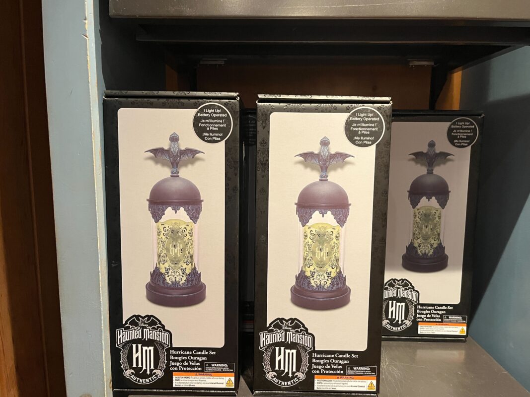 A display shelf featuring three boxed "Haunted Mansion" hurricane candle sets, each box shows an image of the product, which has a gothic-style transparent shade and bat-shaped topper.