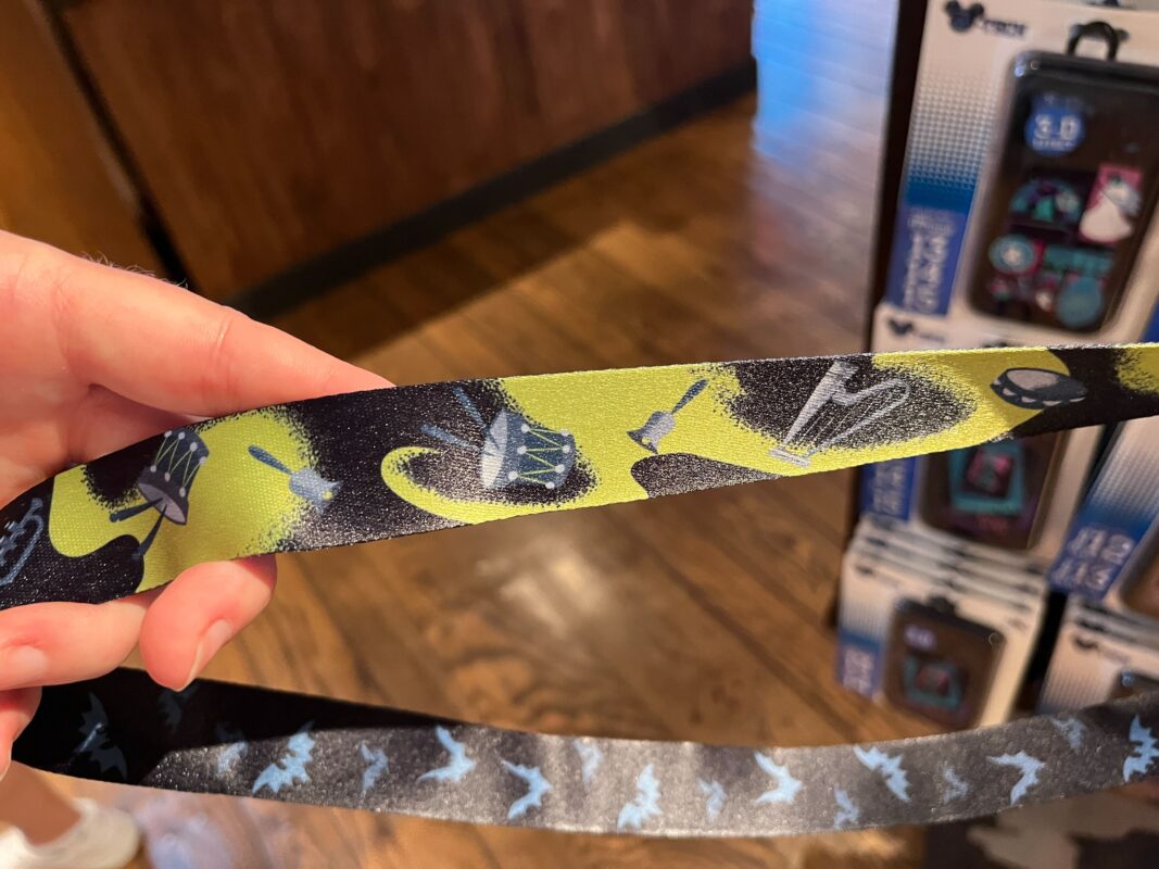 A close-up of a lanyard with green and black sections, featuring illustrations of drums, drumsticks, and musical notes. The background shows a wooden floor and packaged items on display.