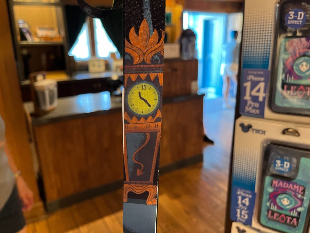 A decorative wooden clock with a yellow clock face, mounted on a shop counter. The shop interior is slightly blurred in the background, with displays of iPhone 14 Pro Max accessories to the right.