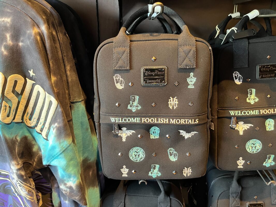 A collection of black backpacks with "Welcome Foolish Mortals" and various small designs displayed on a rack next to a tie-dye shirt with "Imagination" text.