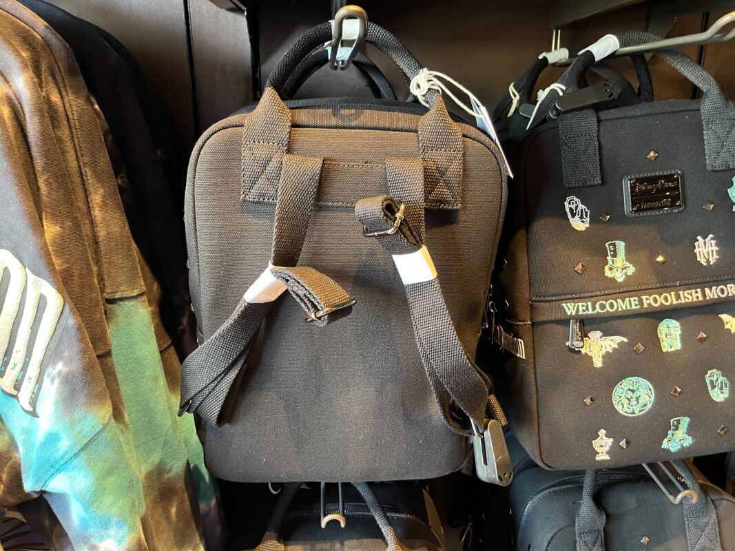 Two black backpacks hang on a display rack. The foreground backpack has adjustable straps with light-colored loop accents, while the other features various green designs and the text "WELCOME FOOLISH MORTALS.