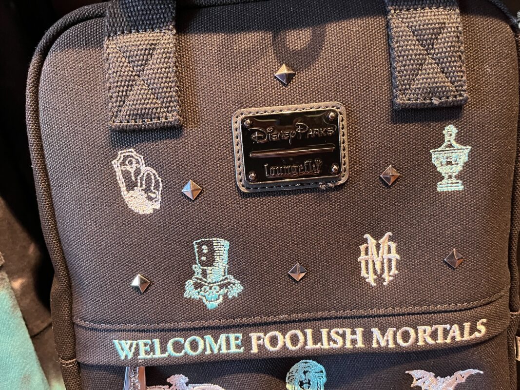A black Disney Parks Loungefly backpack featuring the text "Welcome Foolish Mortals" and various Haunted Mansion-themed designs, including icons of ghosts and a graveyard urn.