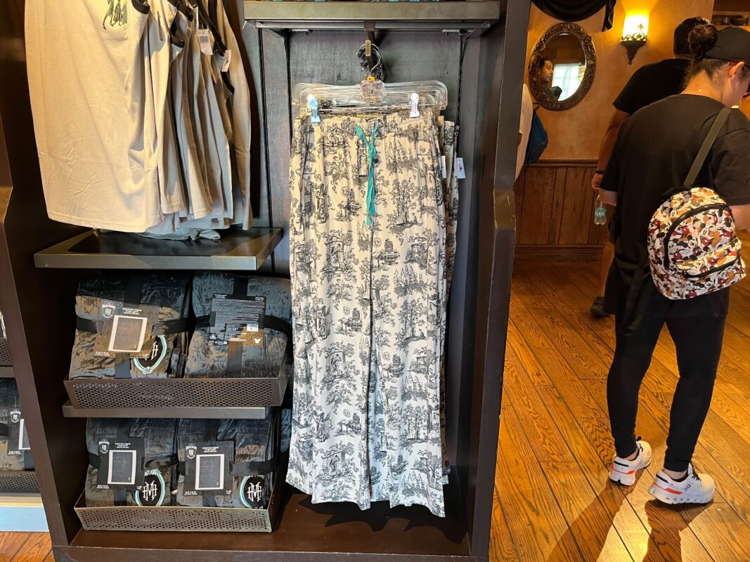 Clothing shop display with pants on hangers, shirts folded on shelves, and a person wearing a floral backpack browsing.