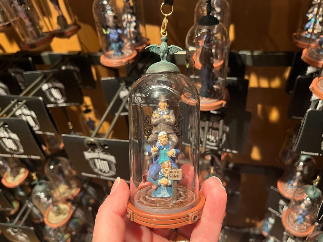A hand holds a decorative figurine encapsulated in a small glass dome, featuring two characters from the Haunted Mansion attraction. Numerous similar figurines are displayed in the background.