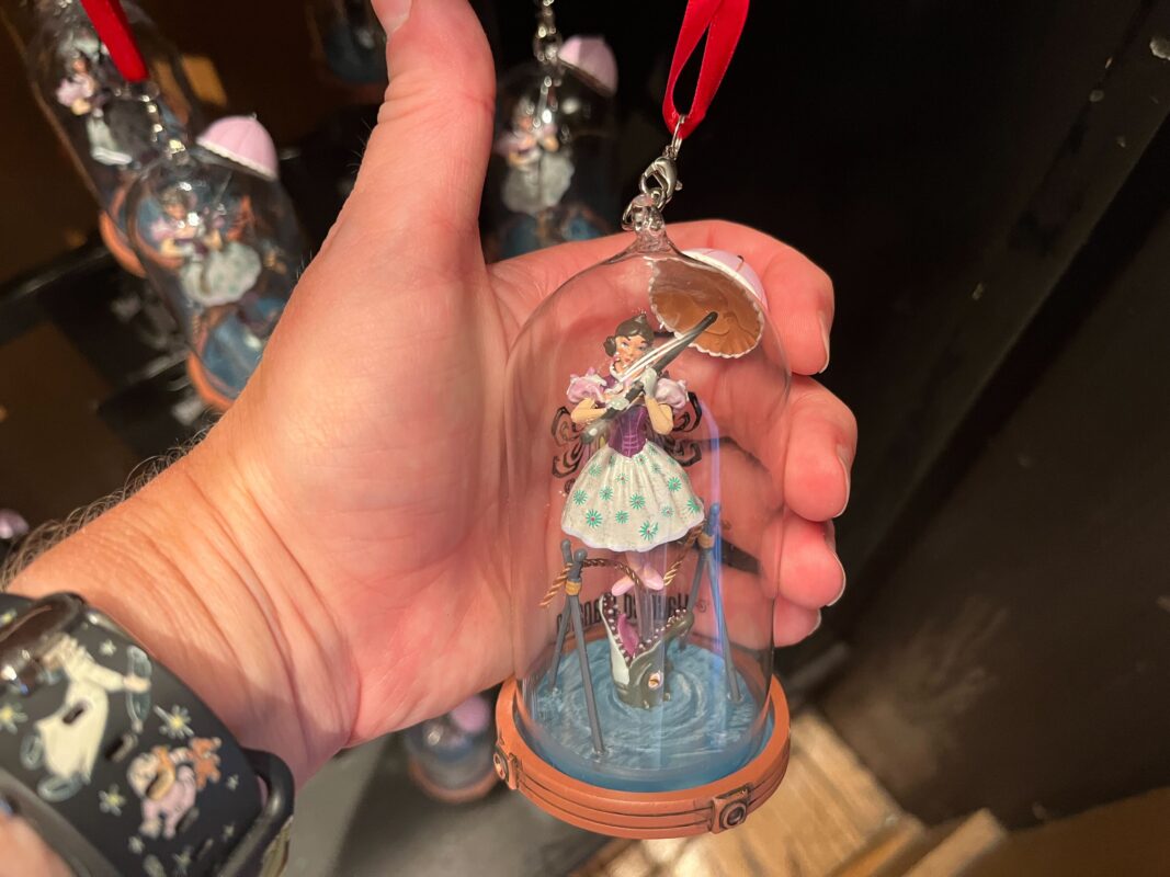 A hand holds a glass dome containing an ornament of a figure dressed in a flowered skirt and wielding a sword on a decorative base. The dome is topped with a red ribbon for hanging.