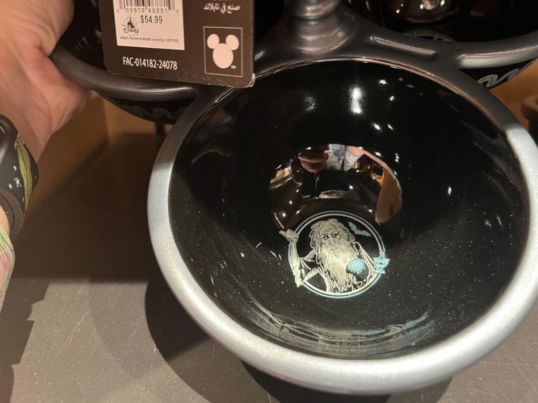 Close-up of a black ceramic bowl with an illustration of a wizard in the center. A price tag showing $54.99 is partially visible hanging from a shelf above. A hand is holding the bowl.