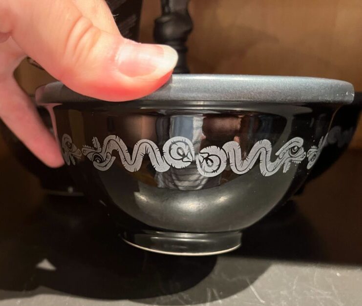 A hand holding a black bowl with a silver wavy pattern near the top edge, against a wooden background.