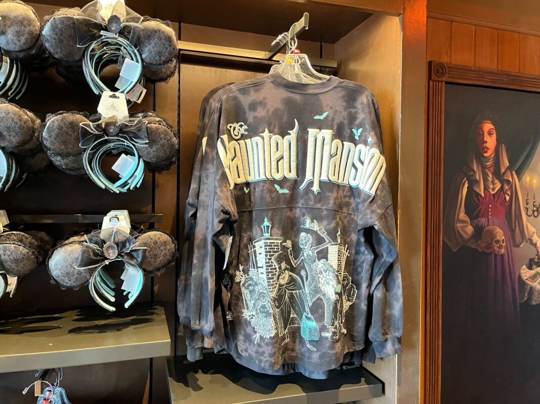 A display of "The Haunted Mansion" merchandise, featuring a sweatshirt with haunted house graphics and identical bat-winged headbands. In the background, there is a painting of a woman holding a skull.