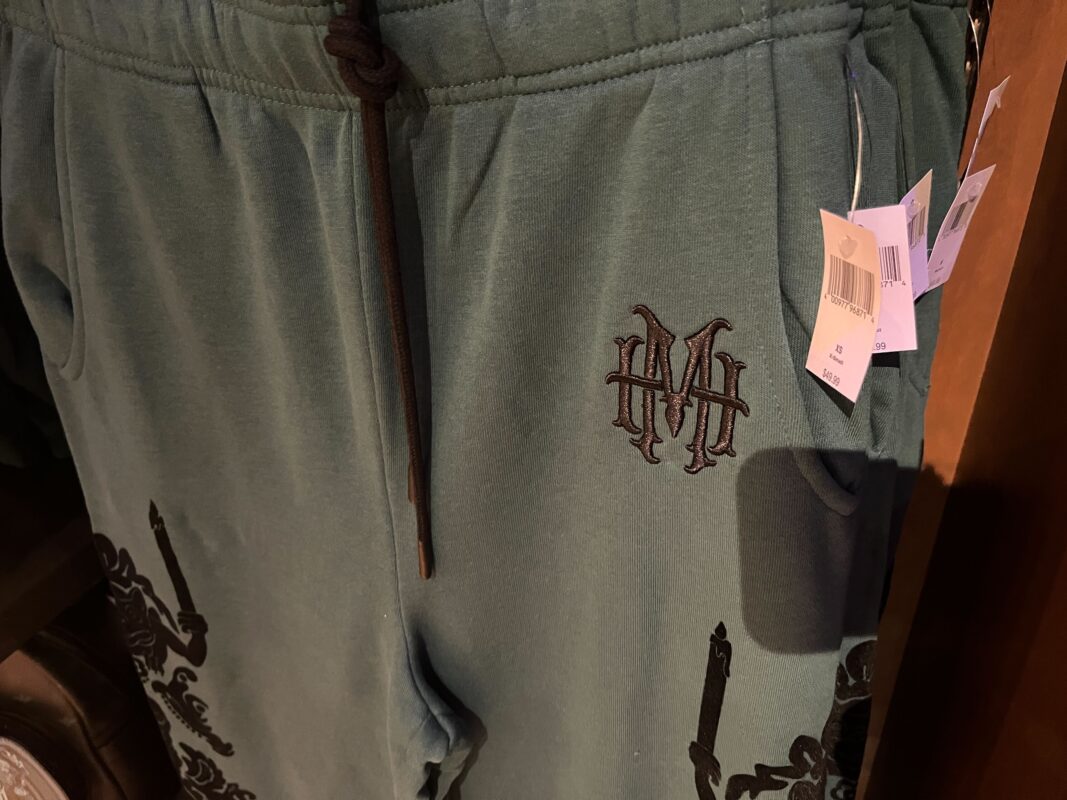 Close-up of green sweatpants with a black "HM" monogram on the left side. Multiple tags are attached to the waistband, and a partial view of a patterned design is visible at the bottom of the pants.