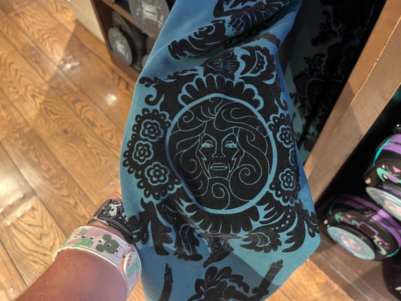 A person holding a blue fabric with black ornate patterns and a central face design. The individual is wearing a wristband on their left wrist.