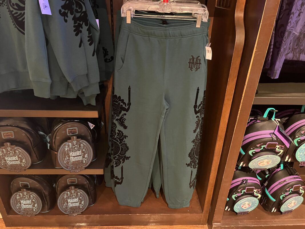 Green sweatpants with a decorative black pattern hang on a display rack. To the left, there are folded sweatshirts, and to the right, round bags with a purple and teal design are on shelves.