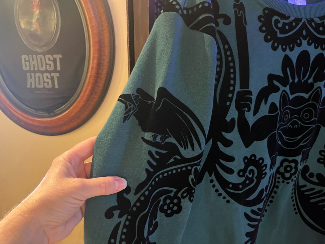 A hand holding a green garment with a black bird and decorative patterns. A framed "Ghost Host" sign is visible in the background.