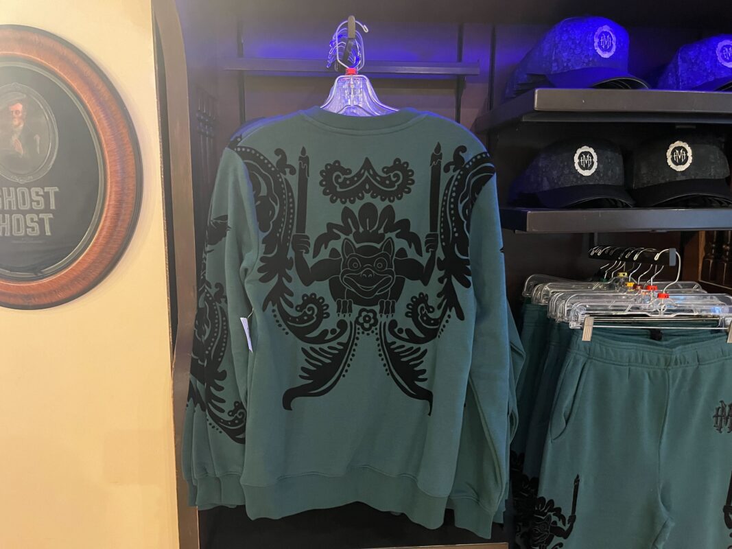 A green sweatshirt with black intricate patterns hangs on a rack, alongside matching green pants and black hats, displayed in a clothing store.