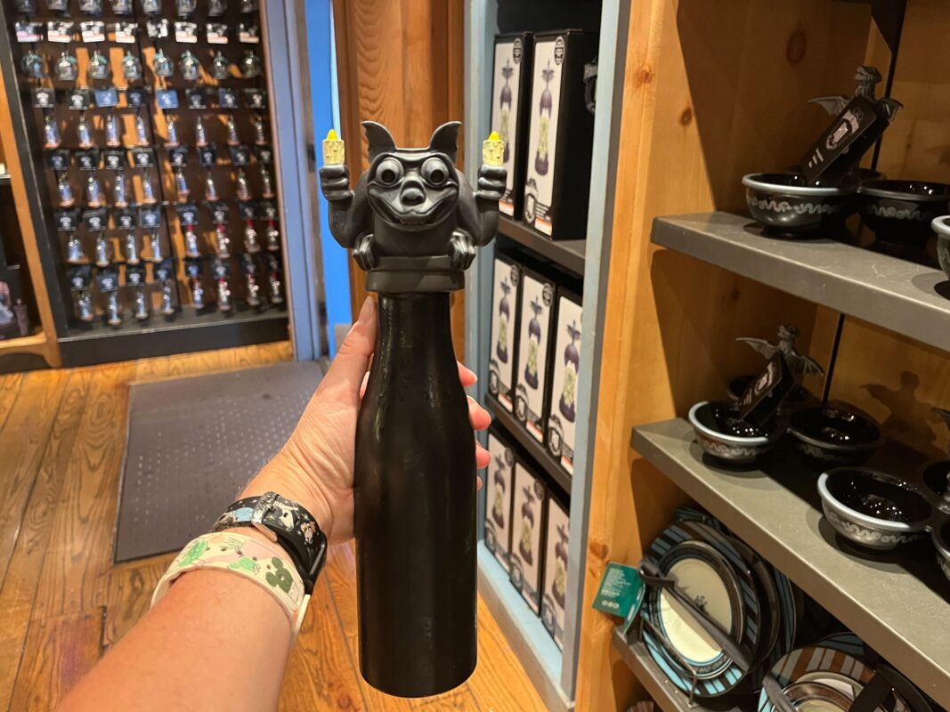 A person holds a black bottle topped with a gargoyle figurine holding what appear to be two candles in a store. Shelves with various items, including similar bottles and ceramic bowls, are in the background.
