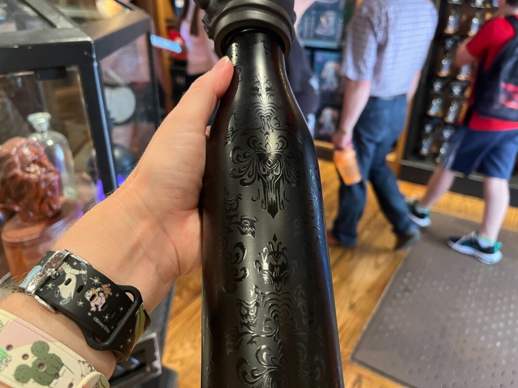 A hand holding a black, patterned bottle inside a store. Shelves with items and people in the background.