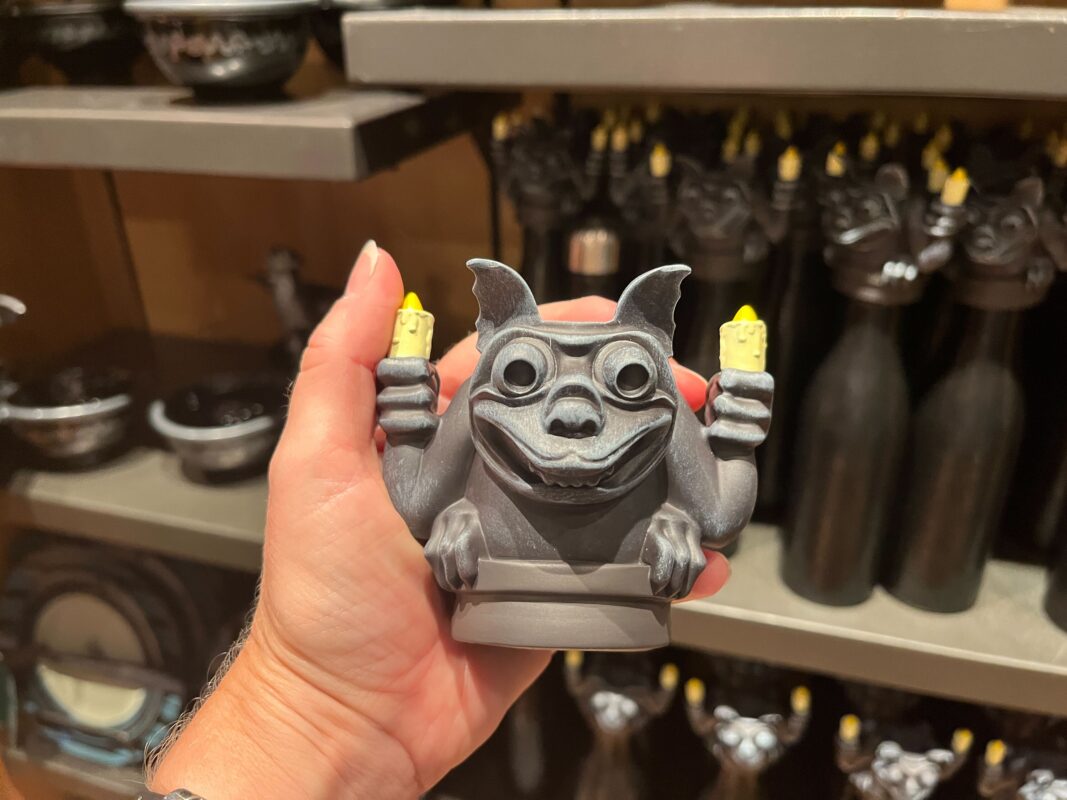 A person holds a small statue of a grey gargoyle with two upraised candles. The background shows shelves with similar items.