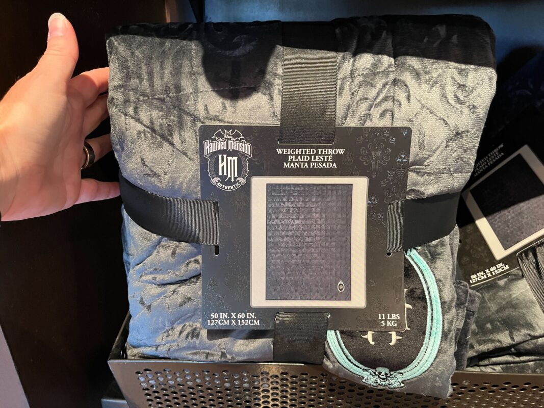 A packaged Haunted Mansion weighted throw blanket with dimensions 50 x 60 inches and weight 12 pounds. The blanket is textured with a dark pattern and secured with a black strap.