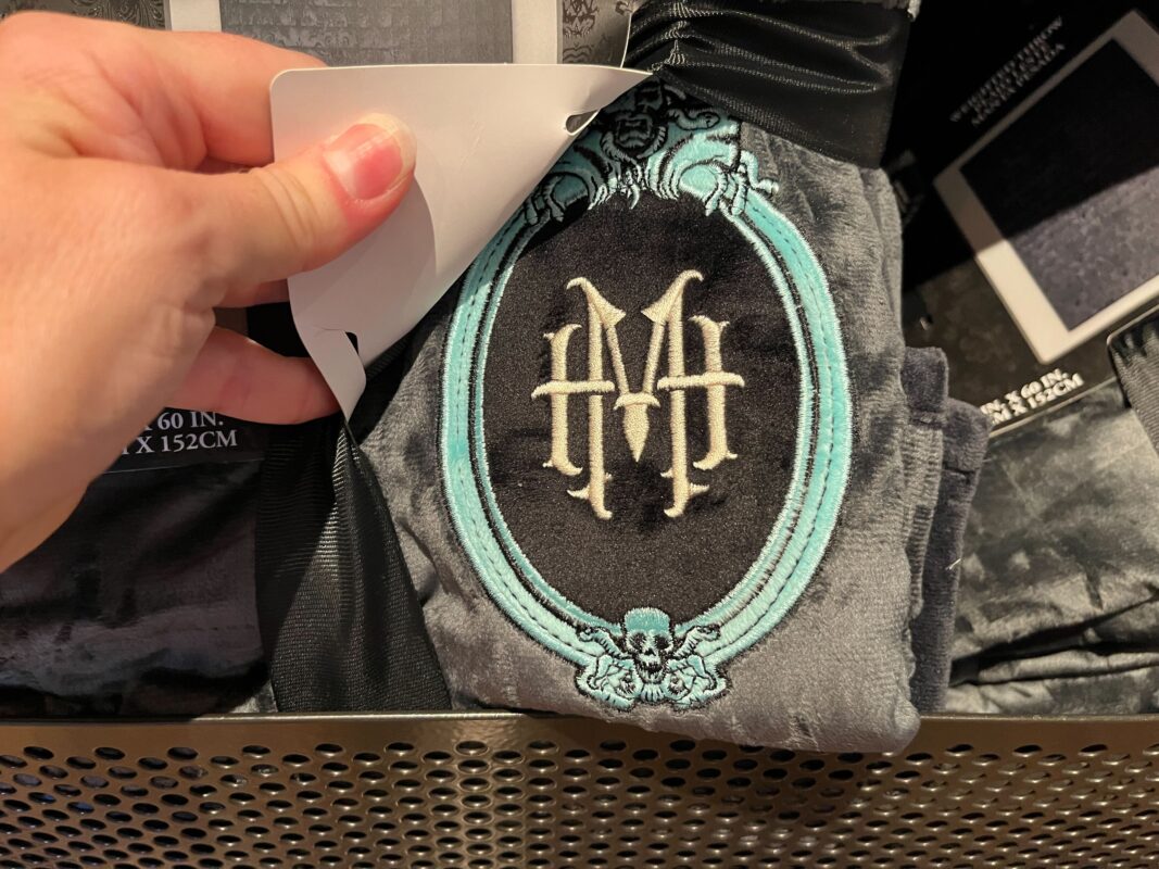 Hand holding a gray fabric with an embroidered insignia in an ornate blue frame, displaying intertwined "H" and "M" letters.