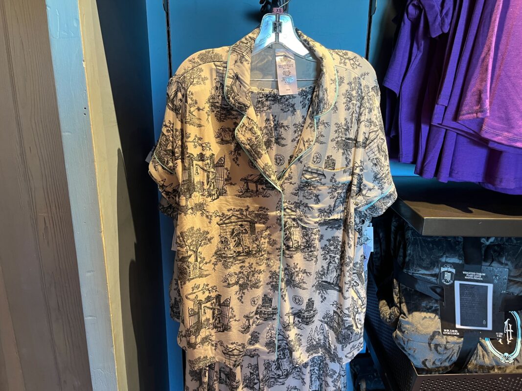 A short-sleeved shirt with a black and white scenic print hangs on a display hook in a store, with purple shirts and other items in the background.