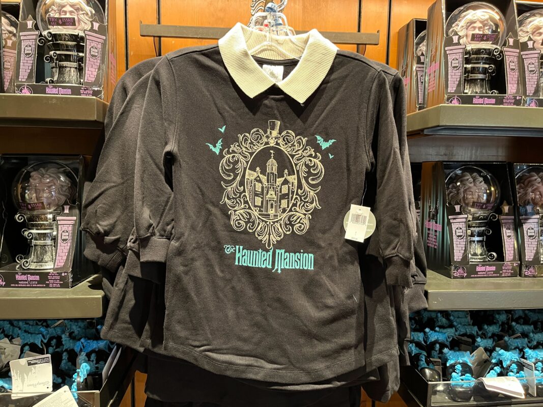 Black shirt with beige collar featuring a "Haunted Mansion" graphic of a house and bats. Displayed on a hanger in a store, surrounded by other merchandise.