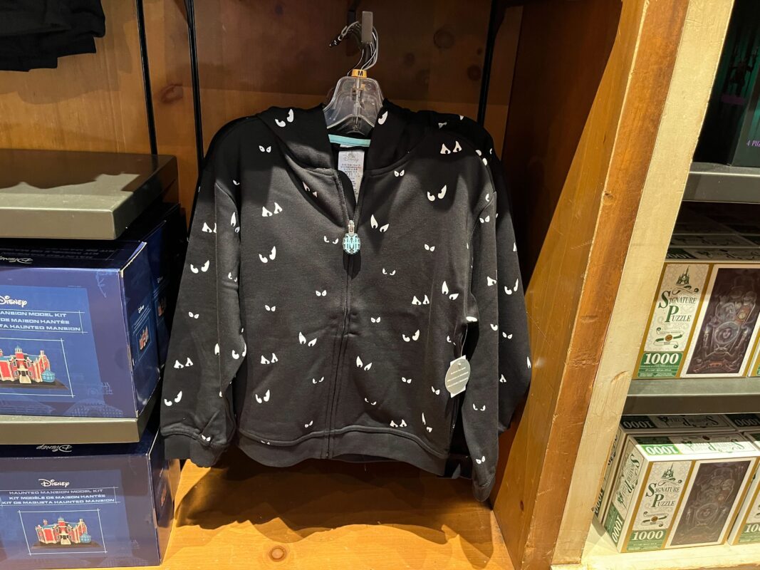 A black hoodie adorned with small white cat face designs is displayed on a wooden shelf in a store. Boxes of various products are arranged on either side.