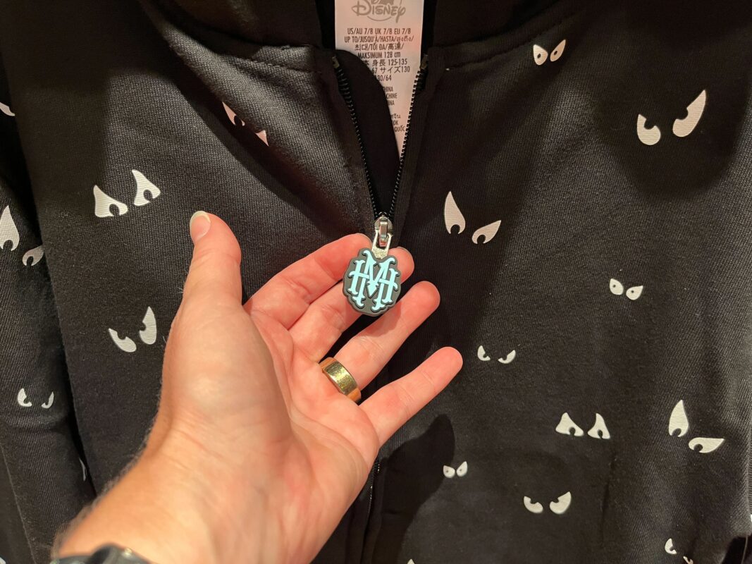 A hand holding the zipper of a black jacket with multiple white eye designs. The zipper pull has a light blue emblem with the letters "HH.