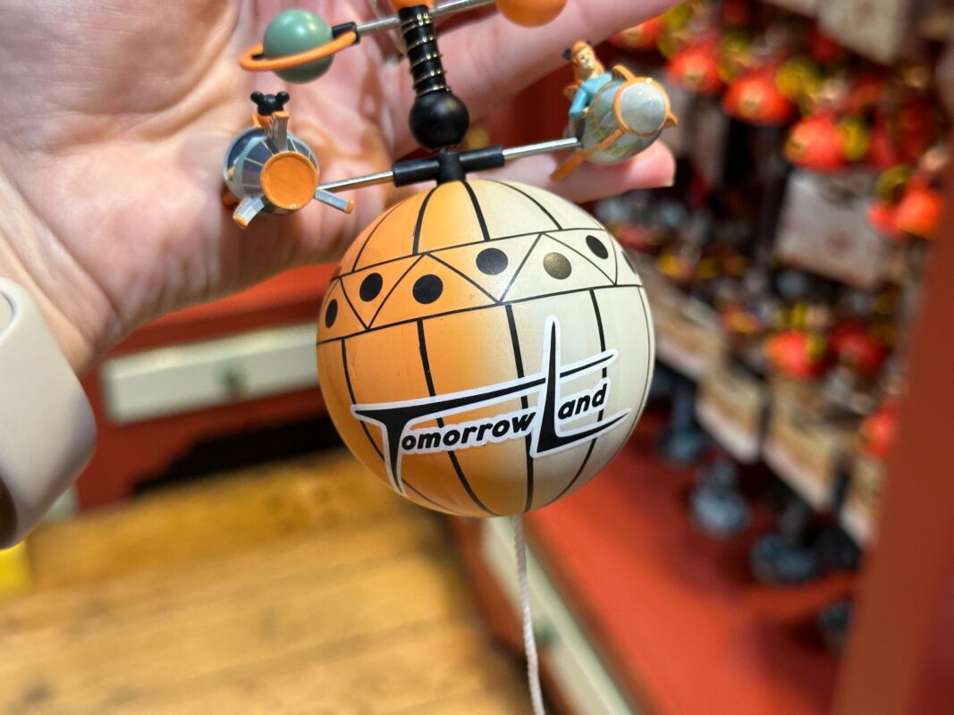 A handheld decorative orb labeled "Tomorrowland," featuring miniature figures and planets arranged around its top, captures the enchanting essence of the Magic Kingdom. This delightful pull string ornament is a whimsical keepsake for any Disney enthusiast.