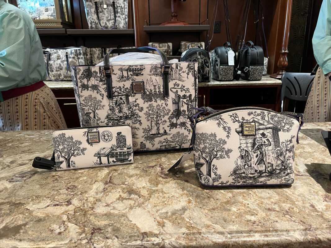A trio of handbags with scenic toile designs displayed on a marble counter. The set includes a wallet, a tote bag, and a small handbag.