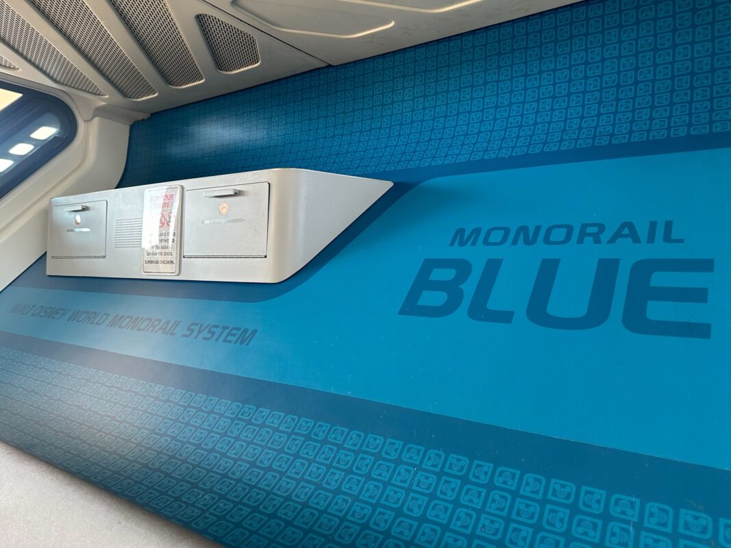 Interior view of a monorail with a blue wall displaying the text "MONORAIL BLUE" and "Walt Disney World Monorail System." The image also shows a wall-mounted shelf with handles.
