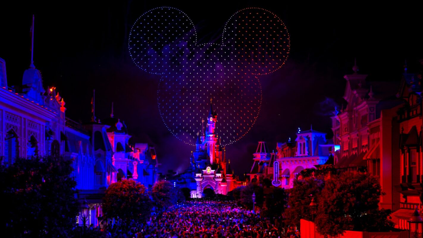 A crowd gathers at a theme park with a colorful castle and a large Mickey Mouse head shape created by drones in the night sky.