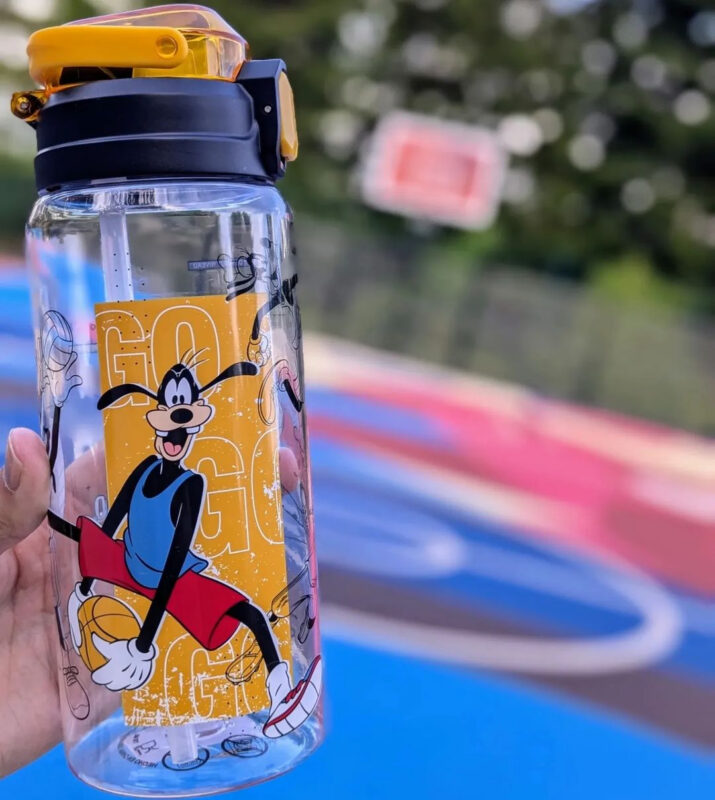 A hand holds a clear water bottle with a cartoon character design in front of a blurred background of a basketball court.