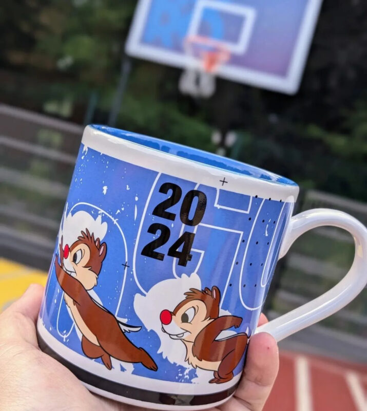 A hand holds a blue mug featuring animated chipmunk characters and the year "2024." A blurred basketball hoop is in the background.