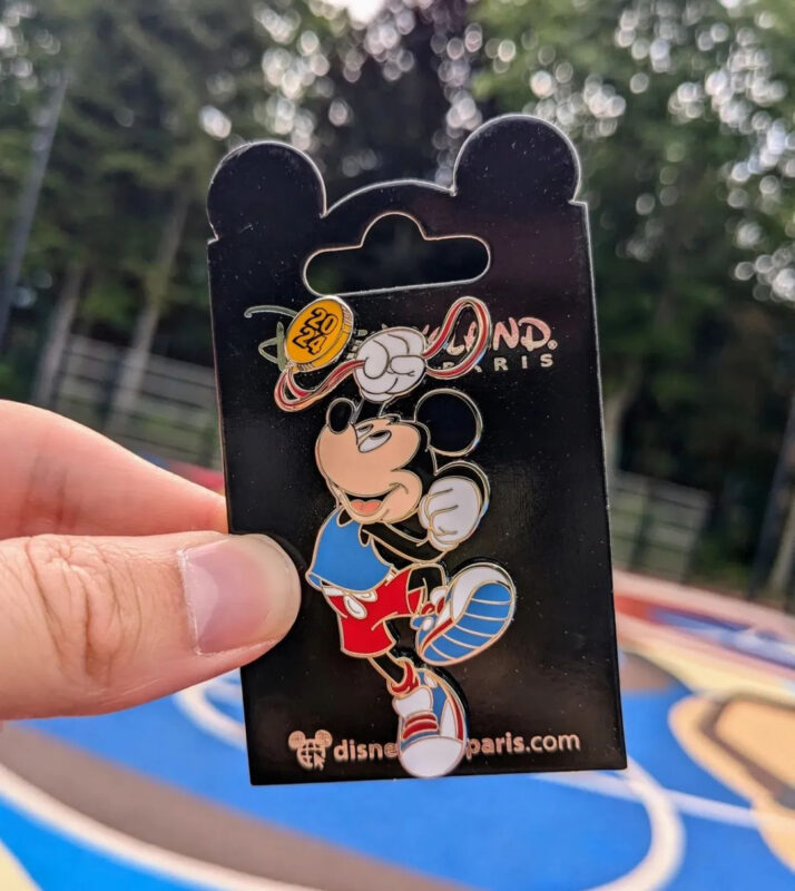 A hand holds a Disneyland Paris enamel pin featuring Mickey Mouse in a red shirt and blue shorts on a card backing, with trees in the blurry background.