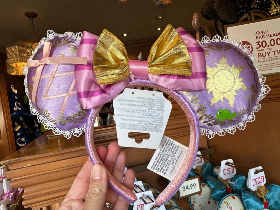 A decorative headband featuring large pink and gold bows with sun and braid designs. Price tag reads 34.99. Displayed on a shelf with other accessories in the background.