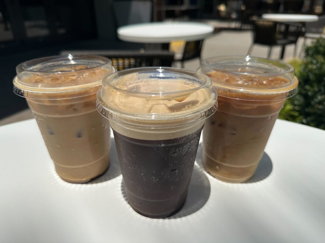 Three plastic cups of iced coffee beverages with lids, one black and two with milk, placed on a white outdoor table in a sunny setting.