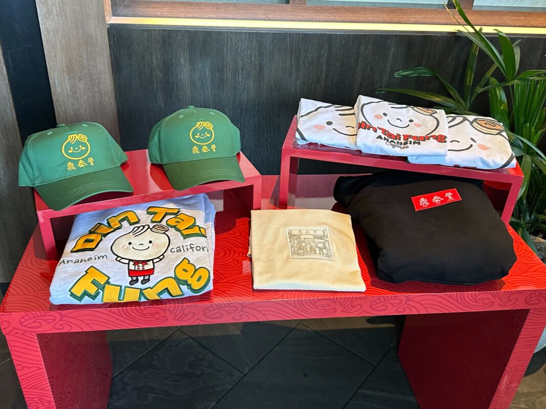 Display of merchandise including green hats with yellow text, folded t-shirts in blue, yellow, and black, placed on red shelves, with a plant visible in the background.