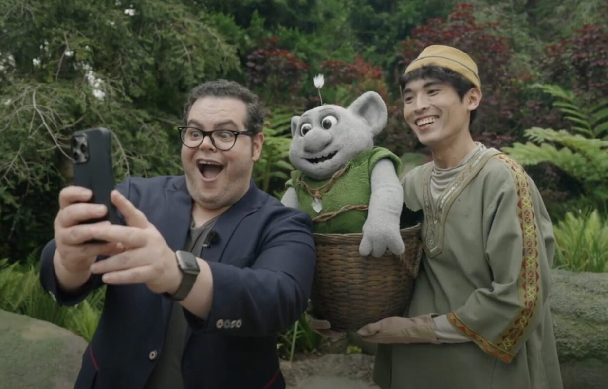 A man with glasses takes a selfie with another man in traditional attire, holding a small gray, costumed character in a basket, all standing in a lush garden at Hong Kong Disneyland’s World of Frozen.