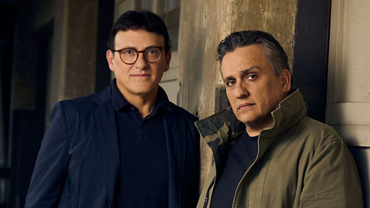 Two men stand in front of a textured wooden background. One wears glasses and a dark blazer, while the other wears a green jacket with the collar turned up. Both look towards the camera.