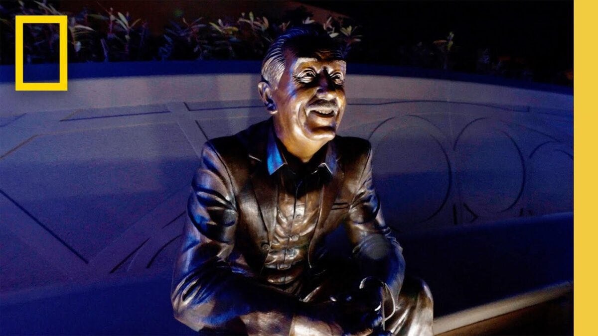 Bronze statue of a smiling man sitting on a bench, illuminated against a blue wall background with plants above.