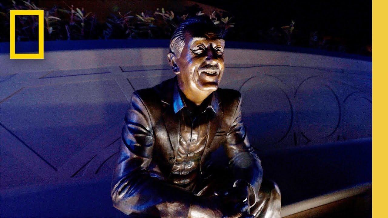 Bronze statue of a smiling man sitting on a bench, illuminated against a blue wall background with plants above.